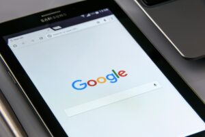 Una tableta Samsung muestra la página de inicio de Google con la barra de búsqueda visible, junto a parte del teclado de un ordenador portátil.