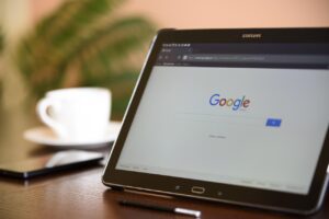 Una tableta Samsung que muestra la página de inicio de la búsqueda de Google está sobre una mesa junto a un smartphone, un lápiz óptico y una taza y un plato blancos. Una planta frondosa aparece borrosa al fondo.