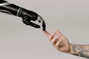 Una mano robótica y una mano humana se tienden la una a la otra, tocándose las yemas de los dedos sobre un fondo liso. El brazo humano tiene tatuajes.