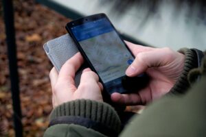 Una persona sostiene un smartphone con una aplicación de mapas y una funda cerrada, mientras está de pie al aire libre con un abrigo.