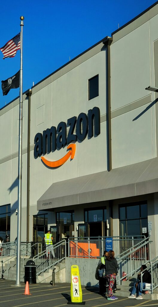 Exterior del almacén de Amazon con un gran logotipo, gente entrando y saliendo, una señal de seguridad en la rampa y una bandera estadounidense en un mástil delante.