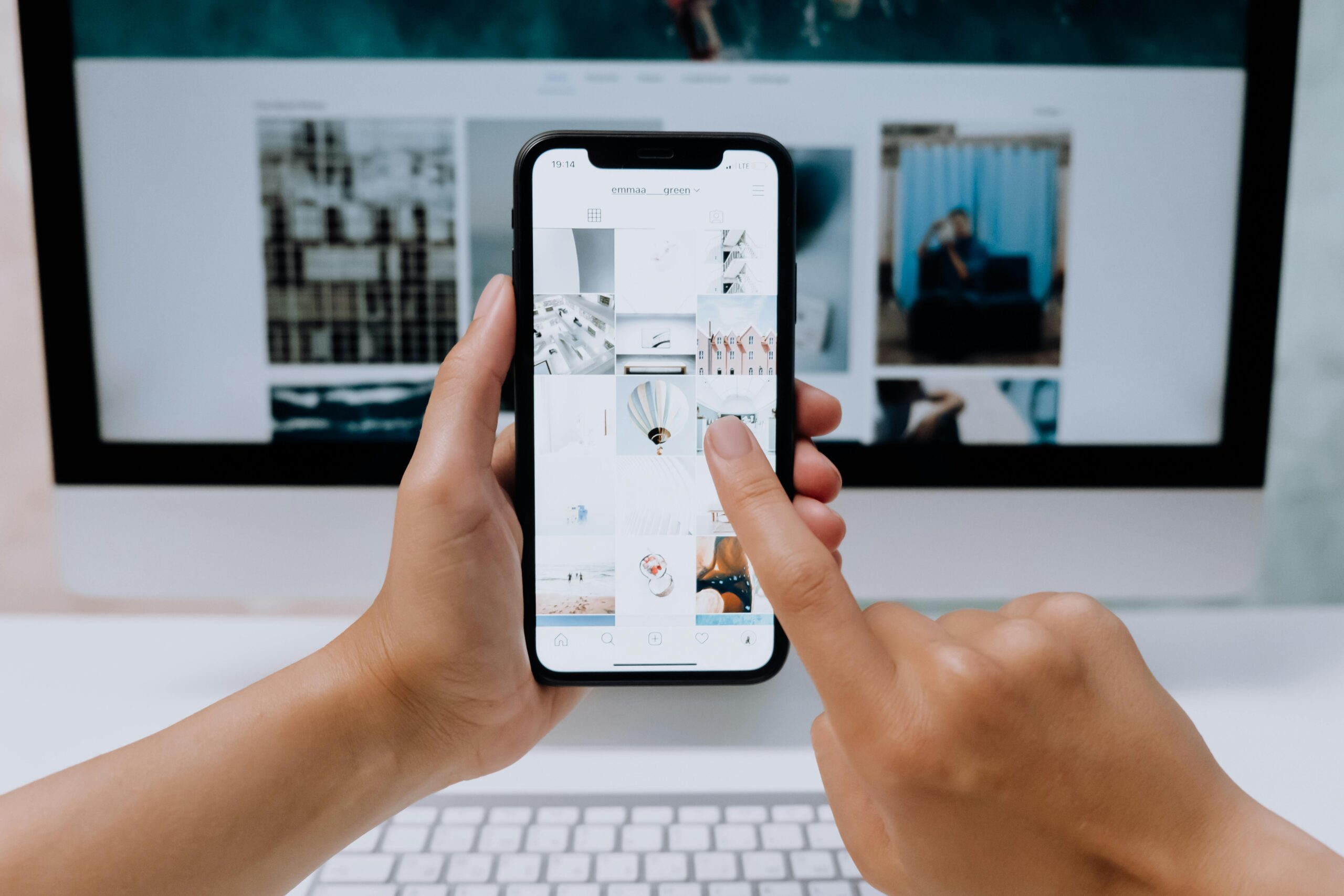 Una persona utiliza un smartphone para navegar por las redes sociales, con el monitor y el teclado de un ordenador en segundo plano.