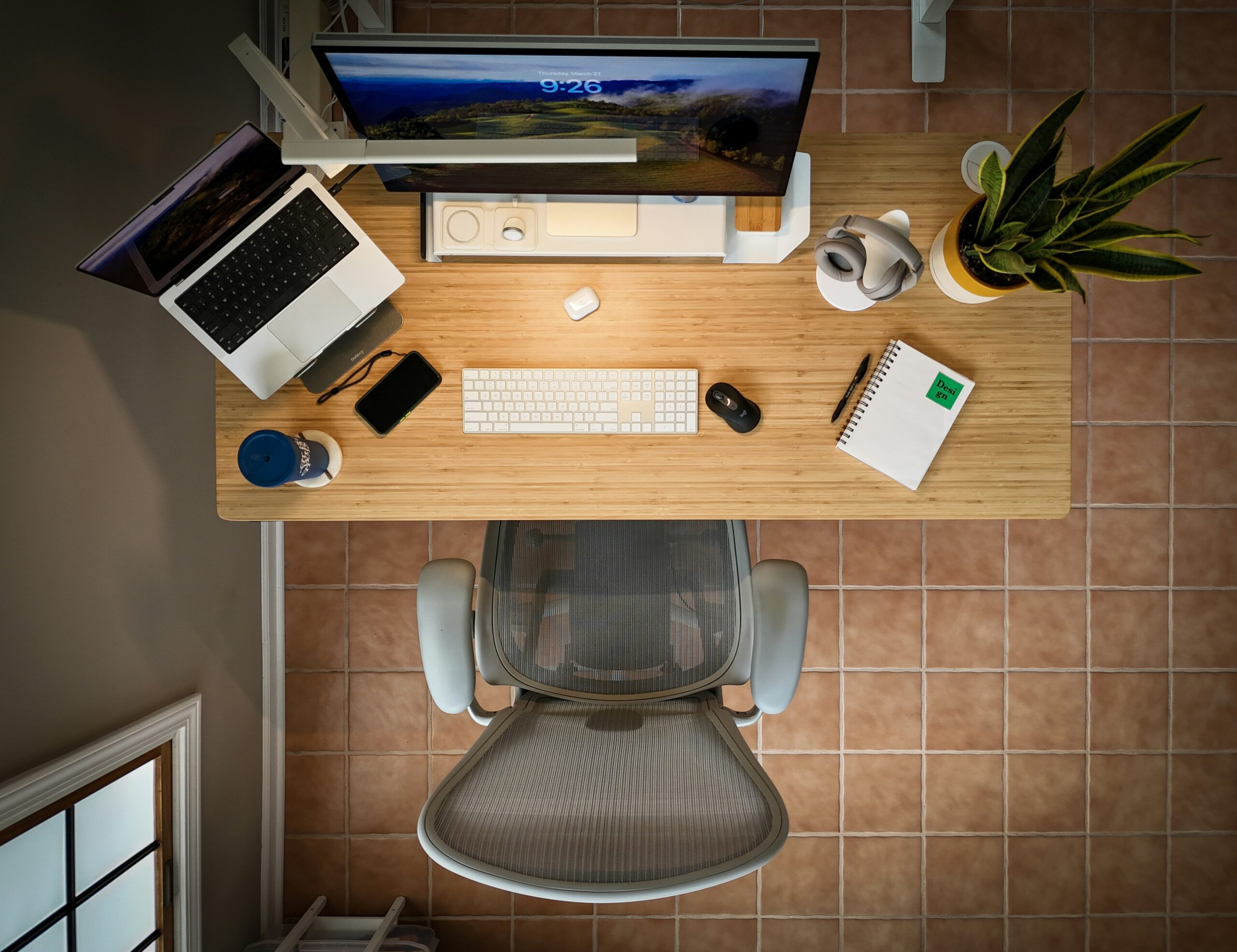 Vista aérea de un escritorio ordenado con un monitor de ordenador, un portátil, un teclado, un ratón, auriculares, un bloc de notas, un teléfono, una taza de café, una planta y una silla de oficina sobre un suelo de baldosas.