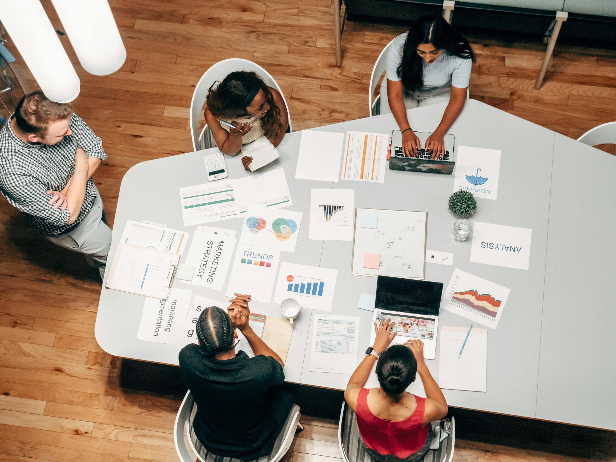 Cuatro personas se sientan a una mesa con ordenadores portátiles y gráficos impresos, mientras una persona permanece de pie cerca; los documentos muestran gráficos, datos e información de marketing.