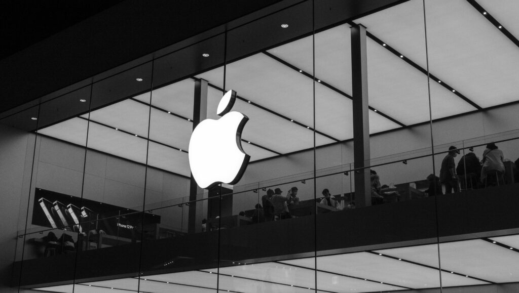 Imagen en blanco y negro de una Apple Store con un gran logotipo de Apple iluminado en la fachada de cristal; se ve gente dentro en un nivel superior.