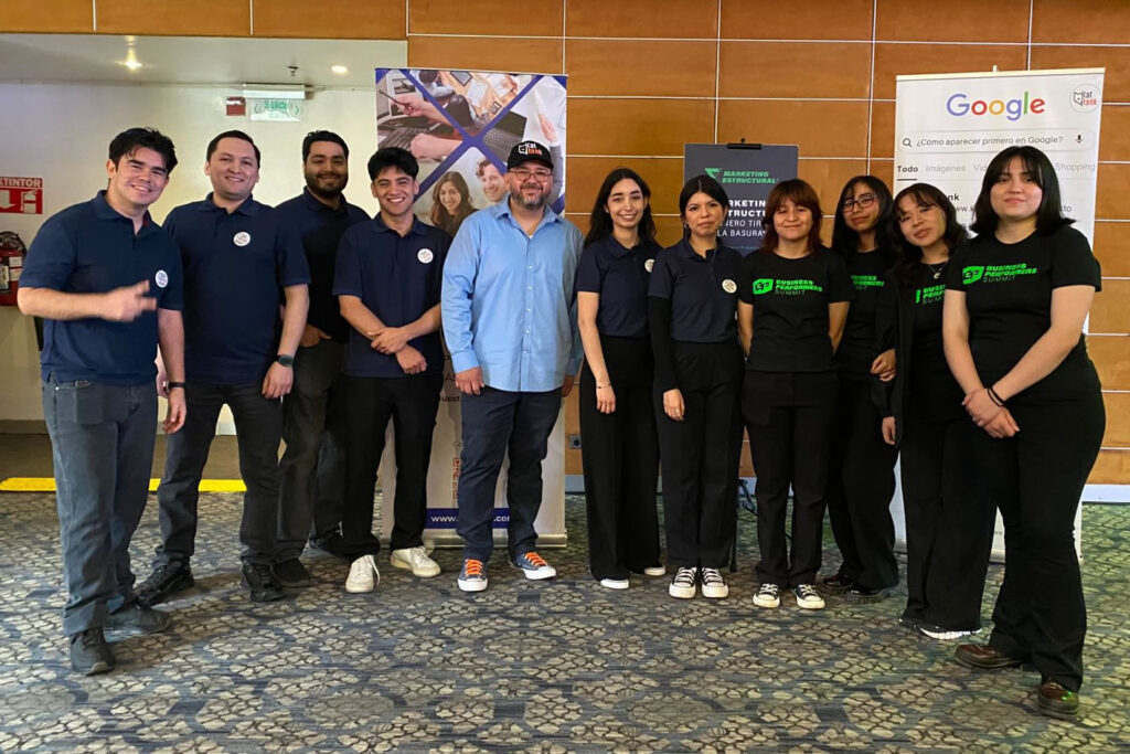 El equipo de KatRank, agencia de SEO en Tijuana, posa en interiores delante de pancartas, algunas con uniformes a juego. El fondo incluye un cartel de Google y un expositor promocional.