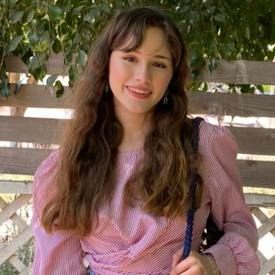 Nosotros 7 Una joven de pelo largo y castaño, con una blusa de rayas rosas, está de pie al aire libre, delante de una valla de madera y vegetación, sonriendo a la cámara.