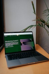 Un portátil sobre una mesa de madera muestra en su pantalla la página de presentación de ChatGPT, con una planta y una pared blanca de fondo.