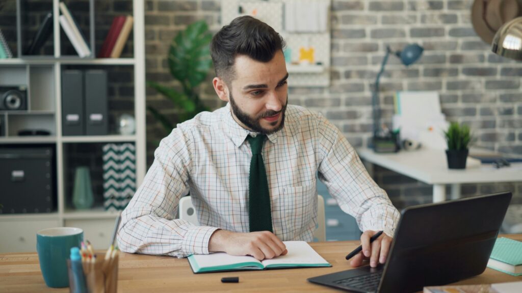 Inicio 17 Un hombre con camisa a cuadros y corbata se sienta en un escritorio con un cuaderno, un bolígrafo y un ordenador portátil en un entorno de oficina moderno, mirando la pantalla del portátil.