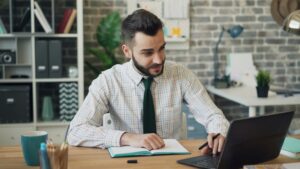 Un hombre con camisa a cuadros y corbata se sienta en un escritorio con un cuaderno, un bolígrafo y un ordenador portátil en un entorno de oficina moderno, mirando la pantalla del portátil.