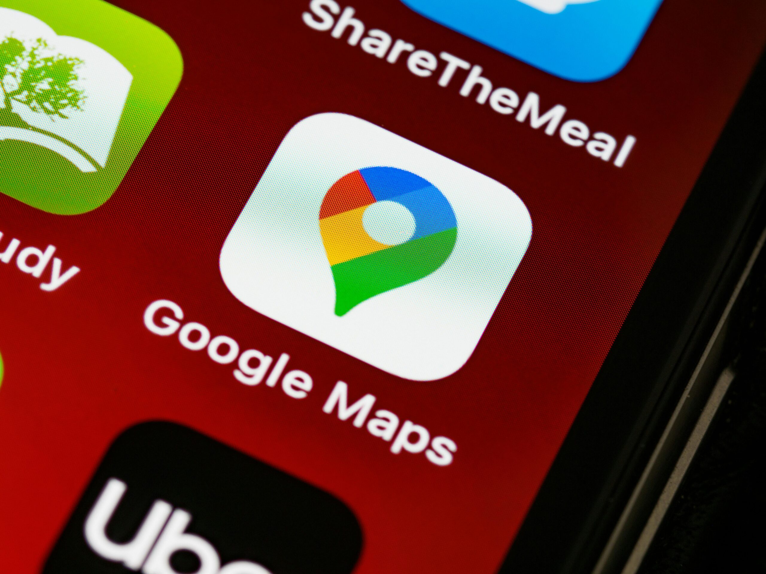 Primer plano de la pantalla de un smartphone que muestra el icono de la aplicación Google Maps con su etiqueta, junto a otros iconos de aplicaciones sobre un fondo rojo.