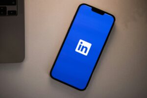 Un smartphone que muestra el logotipo de la aplicación LinkedIn en su pantalla, colocado sobre una superficie de color claro junto a un ordenador portátil.