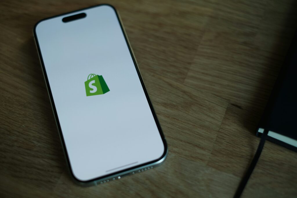 Inicio 14 Un smartphone que muestra el logotipo de Shopify en su pantalla descansa sobre una superficie de madera junto a un cuaderno de tapas negras.