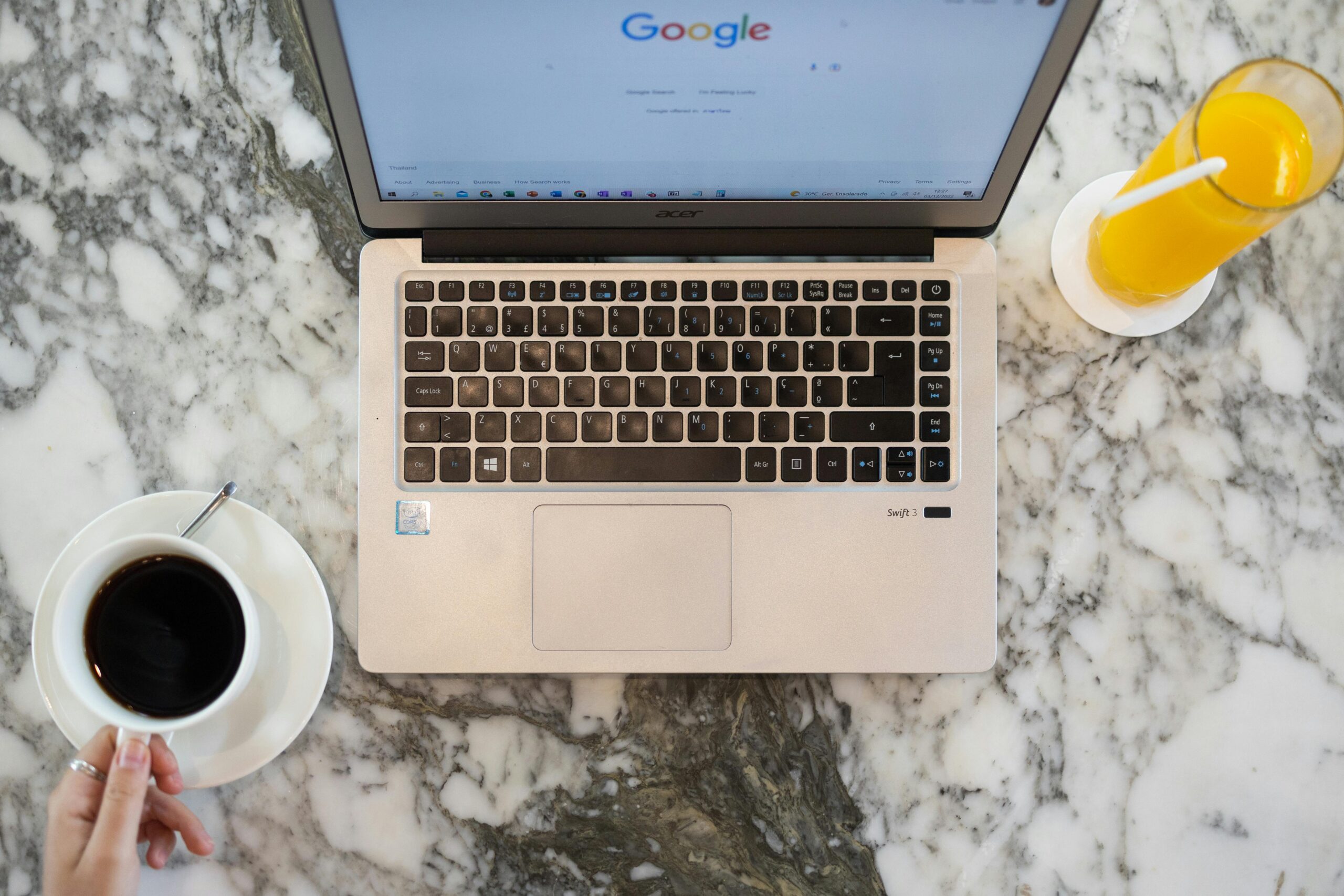Un ordenador portátil con la página de inicio de Google abierta descansa sobre una superficie de mármol, junto a una taza de café solo y un vaso de zumo de naranja.