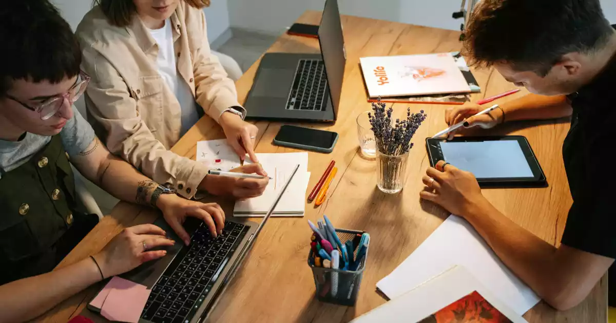Tres personas trabajan en una mesa de madera con ordenadores portátiles, cuadernos y una tableta, colaborando y tomando notas. En la mesa también hay un jarrón con lavanda y varios artículos de papelería.