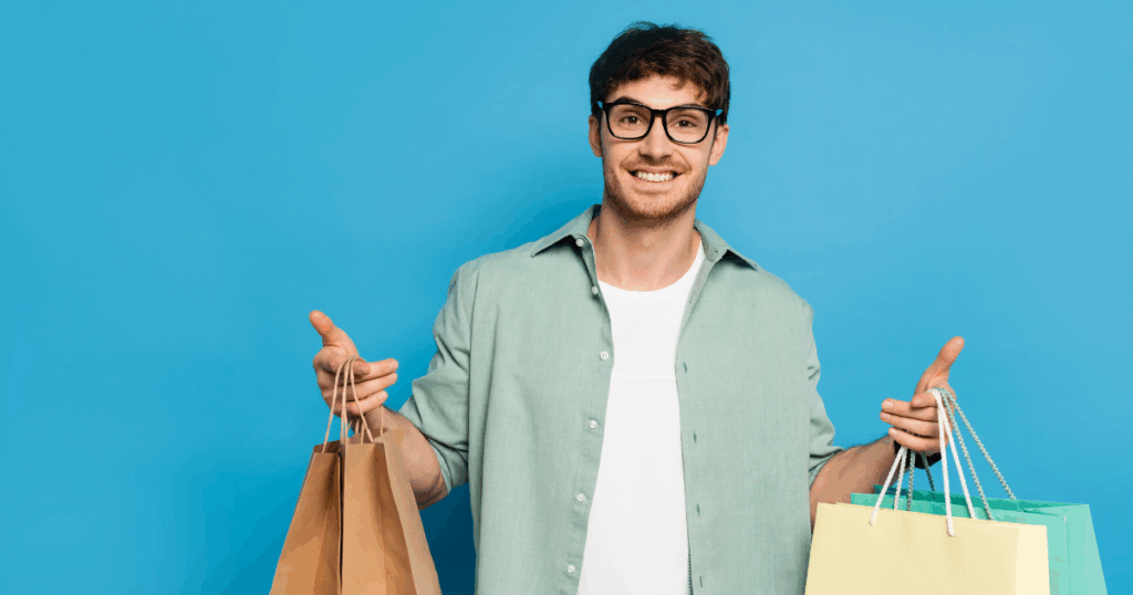 Inicio 10 Un hombre con gafas y camisa verde claro sostiene varias bolsas de la compra y sonríe ante un fondo azul, listo para el fin de año y deseoso de atraer clientes con grandes ofertas.