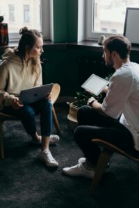 Dos personas sentadas frente a frente en sillas, con un ordenador portátil y una tableta en la mano, conversan en una sala moderna iluminada con luz natural.