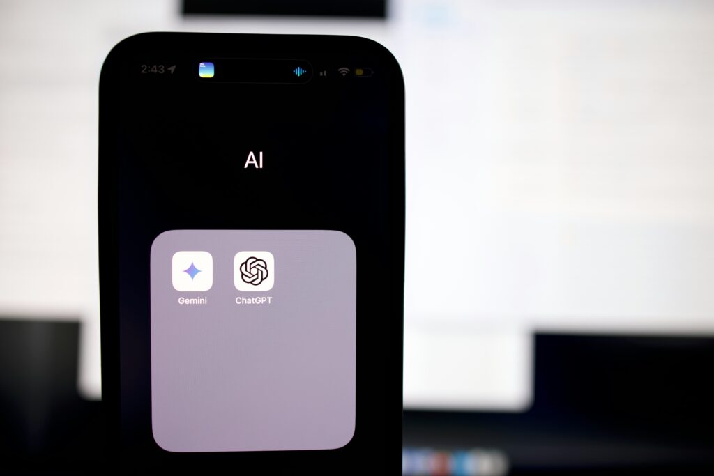 Inicio 17 La pantalla de un smartphone muestra una carpeta con la etiqueta "AI" que contiene los iconos de las aplicaciones Gemini y ChatGPT, con una pantalla de ordenador borrosa de fondo.