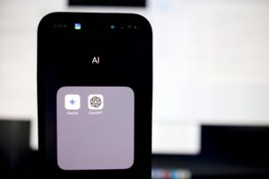 La pantalla de un smartphone muestra una carpeta con la etiqueta "AI" que contiene los iconos de las aplicaciones Gemini y ChatGPT, con una pantalla de ordenador borrosa de fondo.