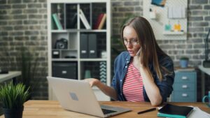 Una mujer con gafas está sentada ante un escritorio, mirando la pantalla de un ordenador portátil con expresión concentrada. Al fondo se ven estanterías con libros y cajas de almacenaje.