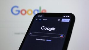 Un smartphone muestra la página de búsqueda de Google en modo oscuro, con la página de inicio de Google también visible borrosa en el fondo.