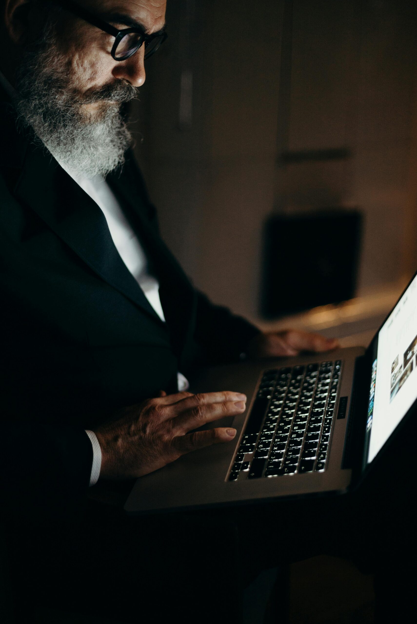 Un hombre con barba y gafas vestido de traje está sentado en una habitación poco iluminada, utilizando un ordenador portátil.