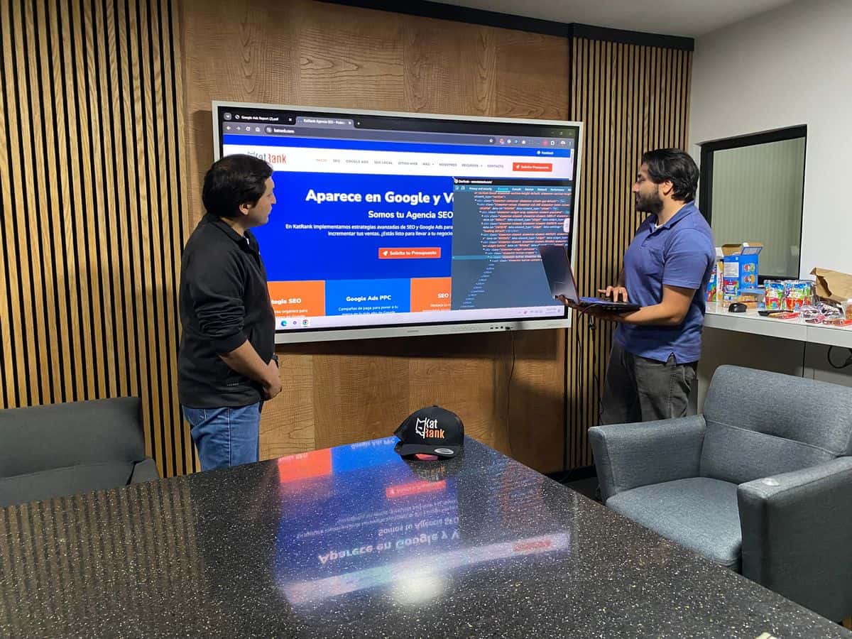 Dos hombres de pie en una oficina mirando a una gran pantalla que muestra un sitio web y su código HTML, discutiendo seo en tijuana; uno sostiene un ordenador portátil, y aperitivos son visibles en una mesa auxiliar.