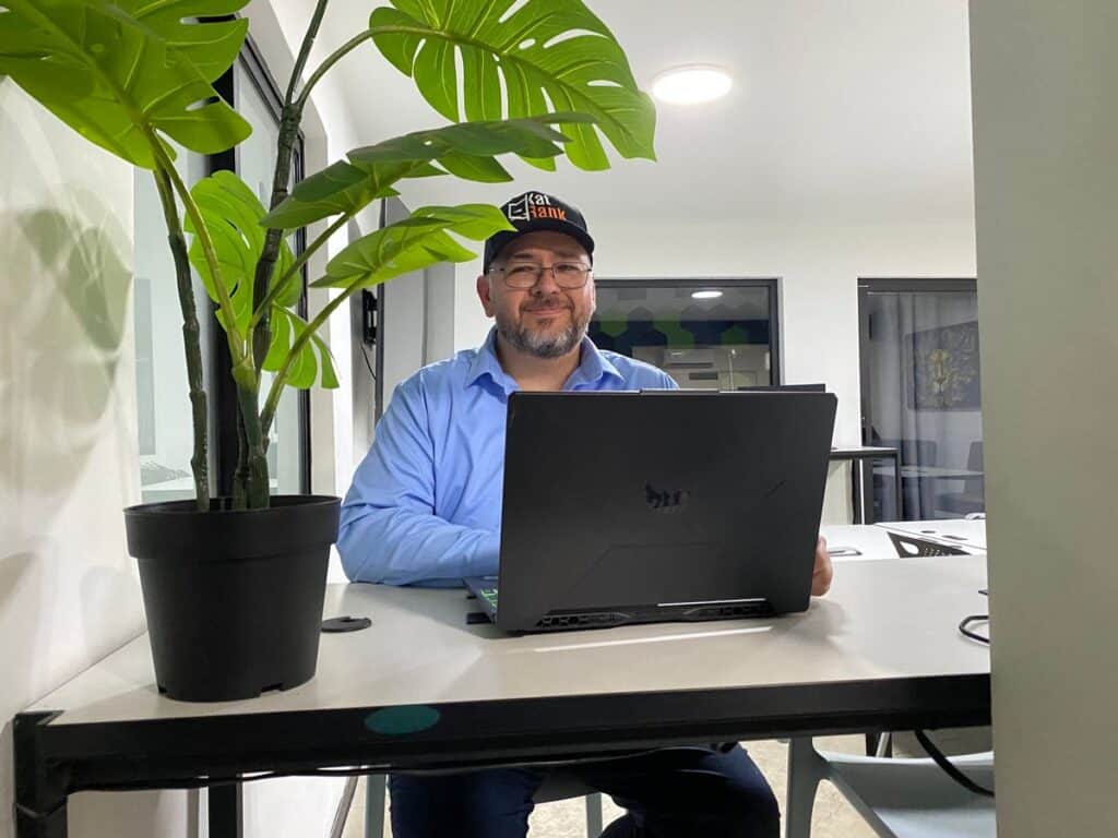 Un hombre con gafas y una gorra se sienta en un escritorio, trabajando en seo en tijuana en su ordenador portátil en un espacio de oficinas modernas con una planta en maceta cerca.