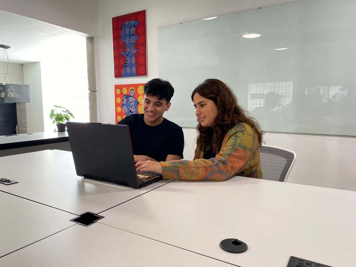 Dos personas sentadas a una mesa en una oficina moderna, mirando un portátil y sonriendo mientras hablan de seo en tijuana. En la pared detrás de ellos cuelgan obras de arte.