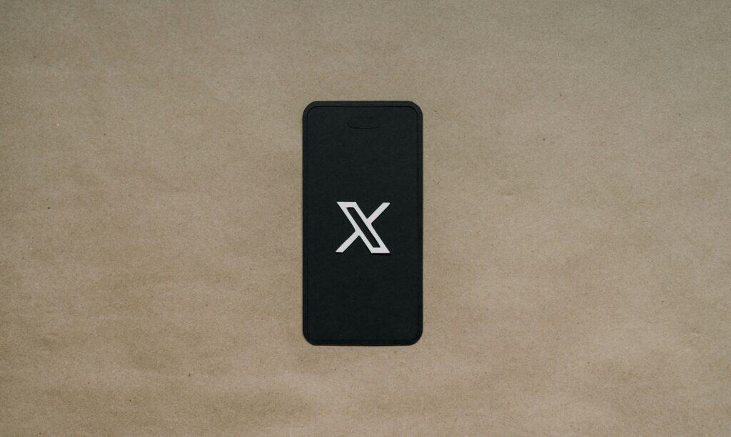 Un smartphone con pantalla negra muestra un logotipo "X" blanco, colocado sobre una superficie texturizada de color marrón claro.