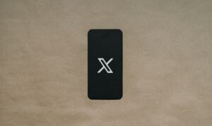Un smartphone con pantalla negra muestra un logotipo "X" blanco, colocado sobre una superficie texturizada de color marrón claro.