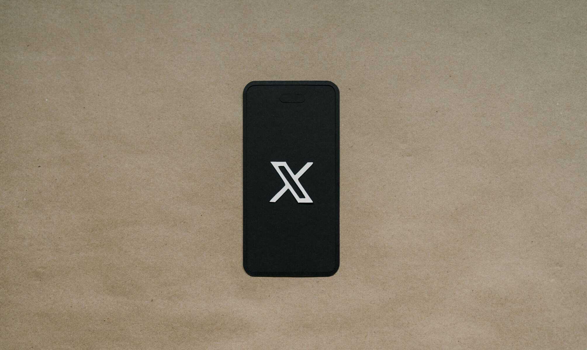 Un smartphone con pantalla negra muestra un logotipo "X" blanco, colocado sobre una superficie texturizada de color marrón claro.