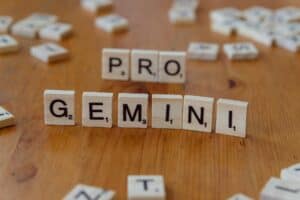 Unas fichas de madera con letras de Scrabble sobre una mesa deletrean "PRO" y "GEMINI", con otras fichas dispersas al fondo.