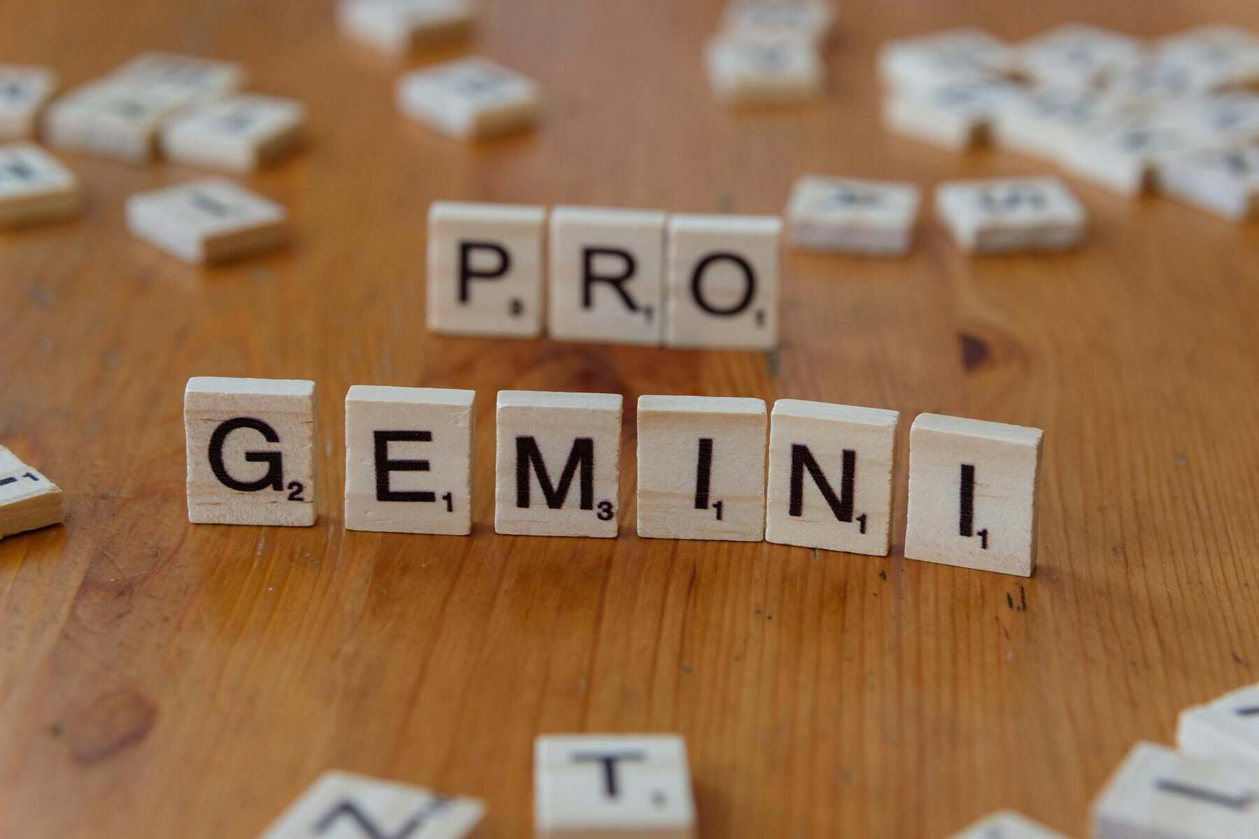 Unas fichas de madera con letras de Scrabble sobre una mesa deletrean "PRO" y "GEMINI", con otras fichas dispersas al fondo.