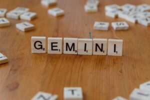 Las fichas de Scrabble de madera que dicen "GEMINI" están dispuestas en fila sobre una superficie de madera, con otras fichas dispersas en el fondo.