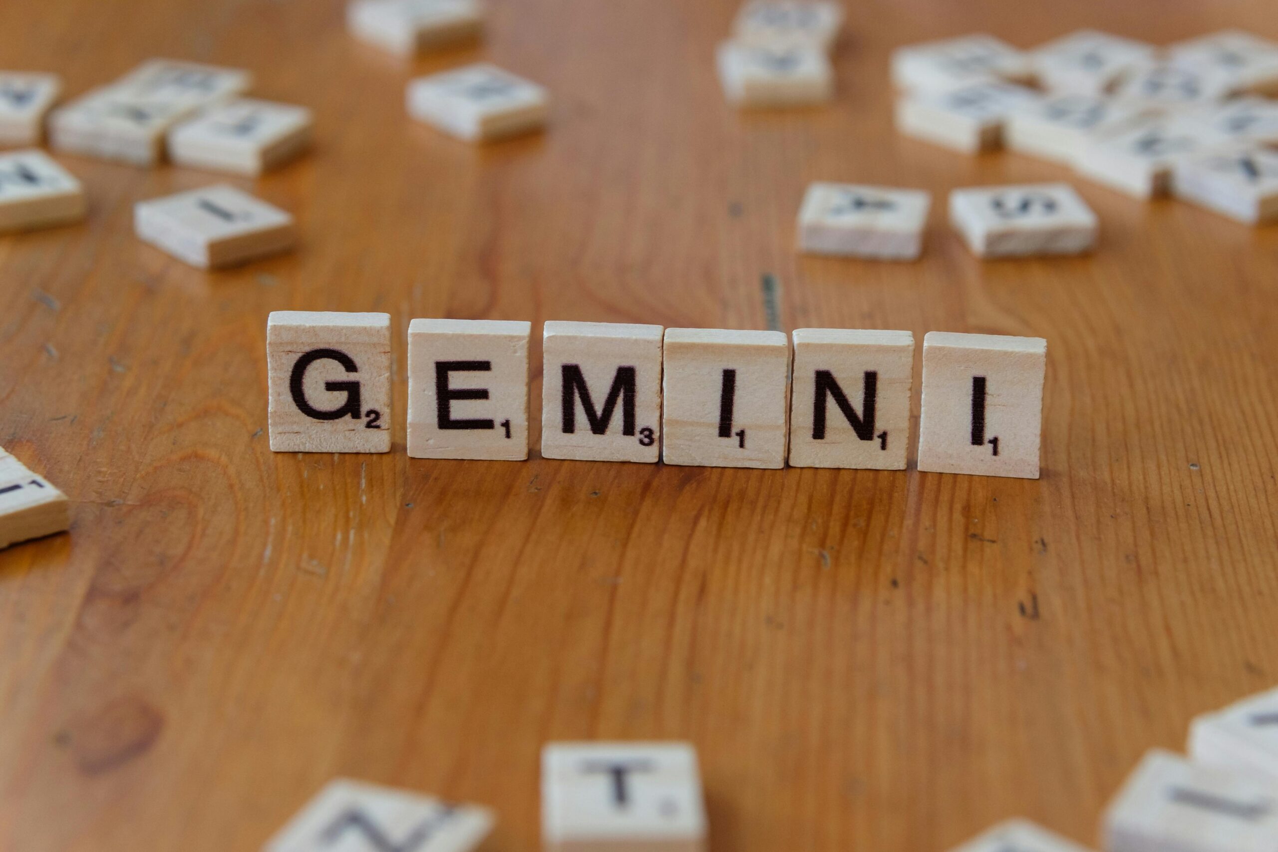 Las fichas de Scrabble de madera que dicen "GEMINI" están dispuestas en fila sobre una superficie de madera, con otras fichas dispersas en el fondo.
