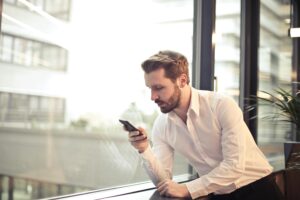 Un hombre con camisa blanca se apoya en el alféizar de una ventana interior, mirando su smartphone.