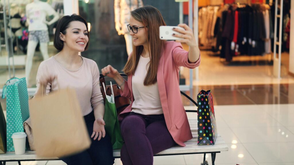 Inicio 14 Dos mujeres sentadas en un banco de un centro comercial, rodeadas de bolsas de la compra, se toman una selfie juntas.