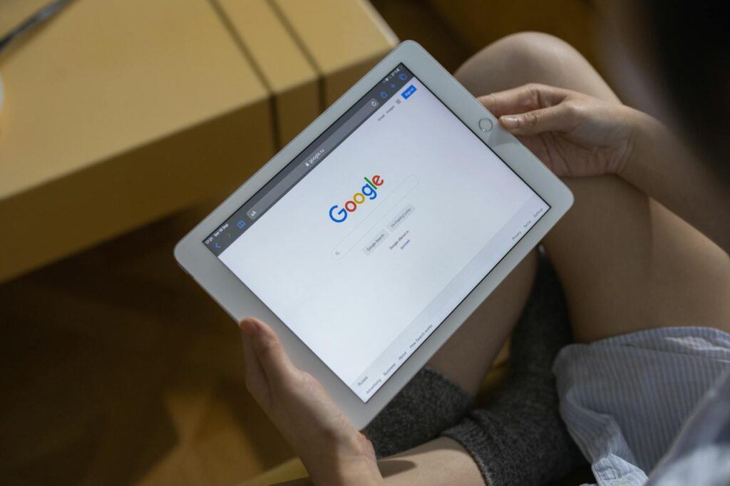Persona que sostiene una tableta blanca con la página de inicio de búsqueda de Google abierta en la pantalla, sentada con las piernas cruzadas en un interior.