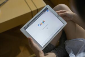 Persona que sostiene una tableta blanca con la página de inicio de búsqueda de Google abierta en la pantalla, sentada con las piernas cruzadas en un interior.