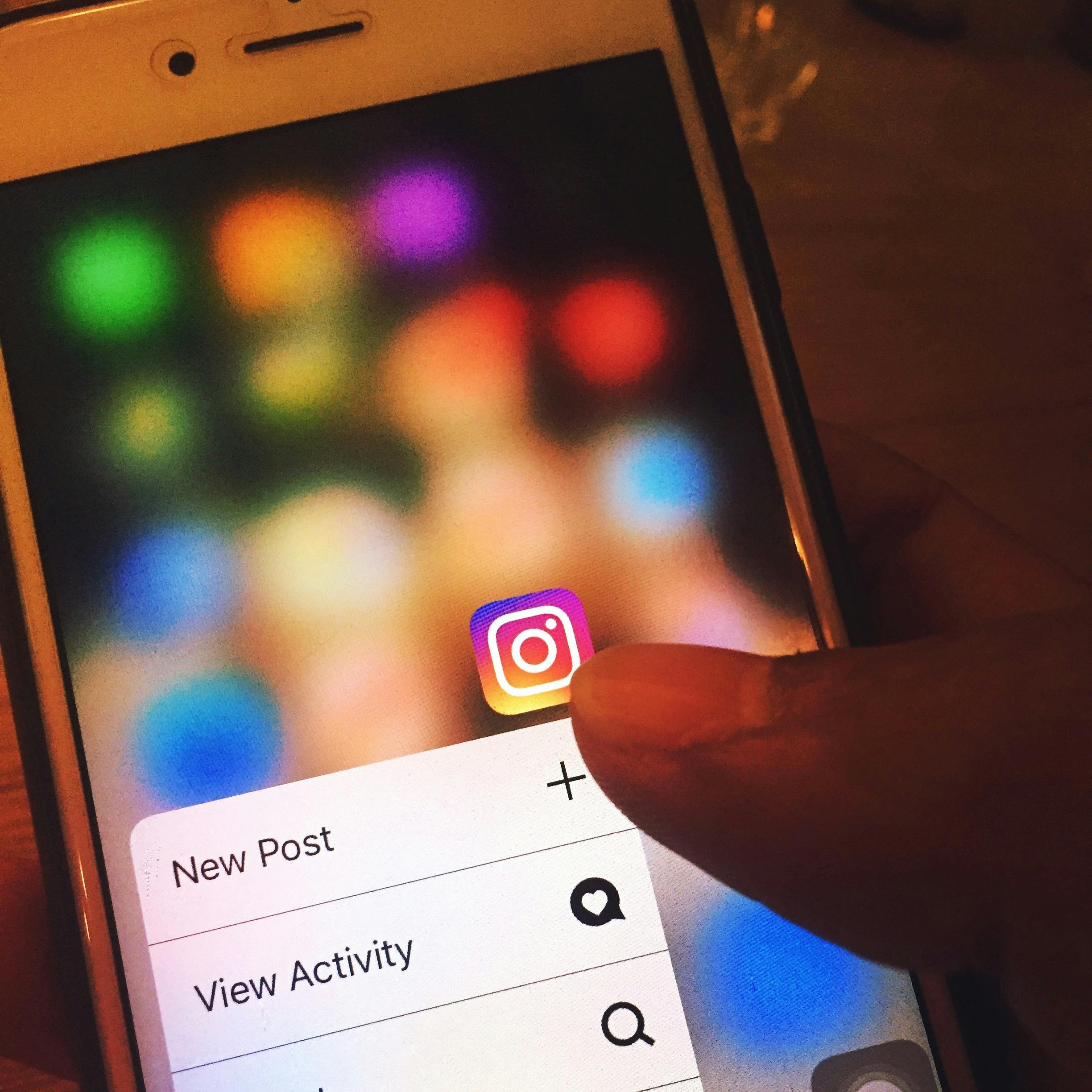 Una mano sujeta un smartphone que muestra el icono de la aplicación Instagram con las opciones "Nueva publicación" y "Ver actividad" en la pantalla.